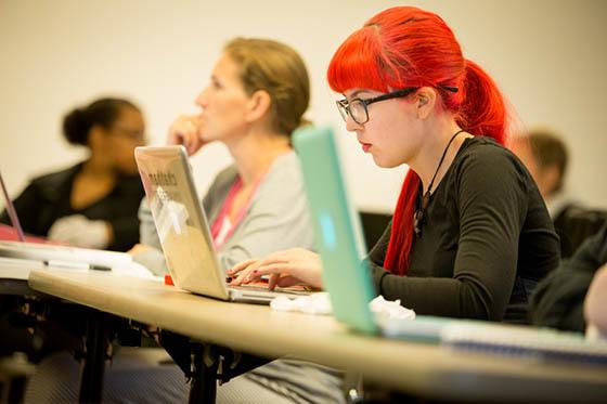 Photo of a student in glasses with bright red hair, working on her laptop in a Chatham University lecture hall