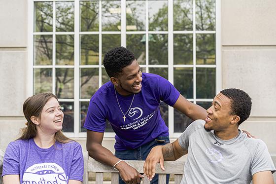 Photo of three Chatham University students sitting outside on a bench laughing and smiling together. 